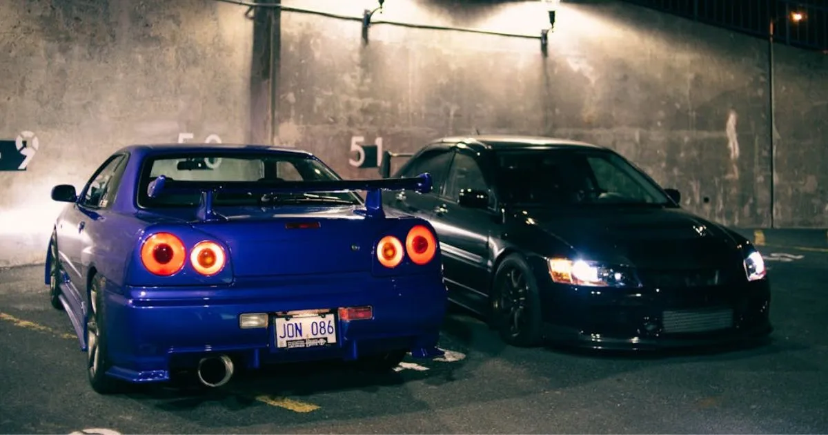 A Black And A Blue Car Parked In A Secure Garage With Their Lights On